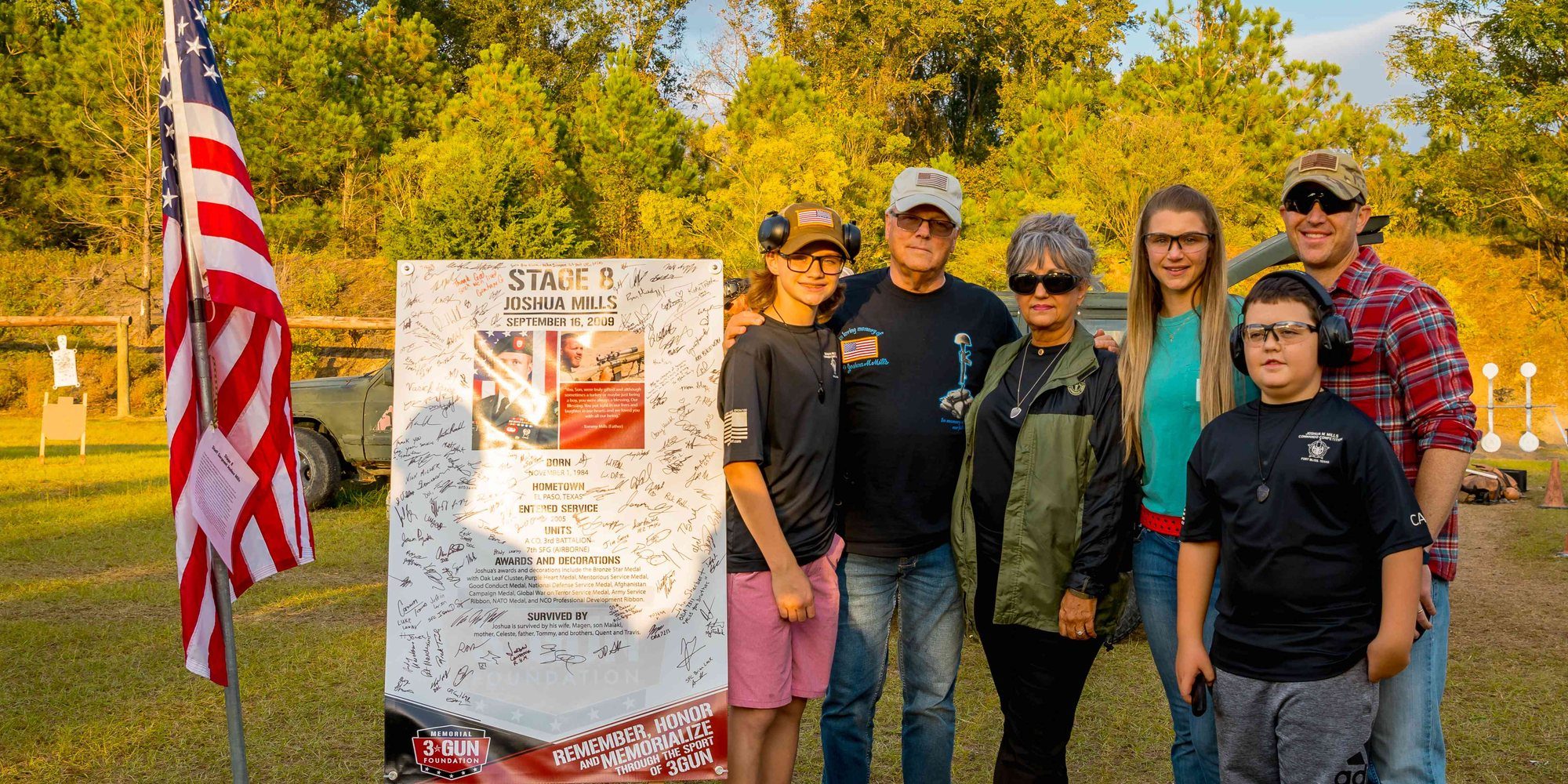 A Gold Star and Surviving family gathers at their honoree's signed stage sign and the American flag at the 2022 Memorial 3 Gun